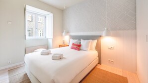 A neatly made bed with white linens and rolled towels, a window with a view of buildings, and a patterned wall.