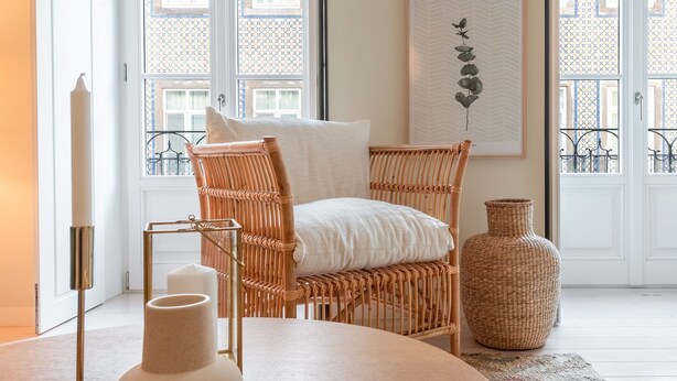 A wicker chair with a white cushion, a round table with a vase, and a woven vase on the floor.