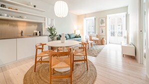 A modern kitchen with white cabinets, a round dining table with wooden chairs, a hanging light fixture, and a view of the living area with a sofa and a plant.