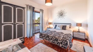 Une chambre avec un grand lit, deux tables de chevet, une commode, un élément décoratif mural et une vue sur l’extérieur par une fenêtre.