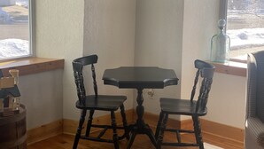 A corner room with a wooden table and two chairs, a window with a view of snow, and a painting on the wall.
