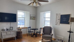 A living room with a TV, wooden flooring, a ceiling fan, a dining table with chairs, a sofa, and a patterned rug.