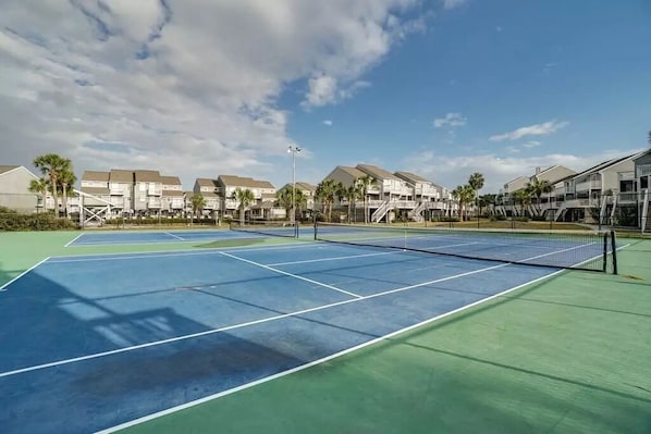 A tennis court with a net, surrounded by buildings with balconies and palm trees.