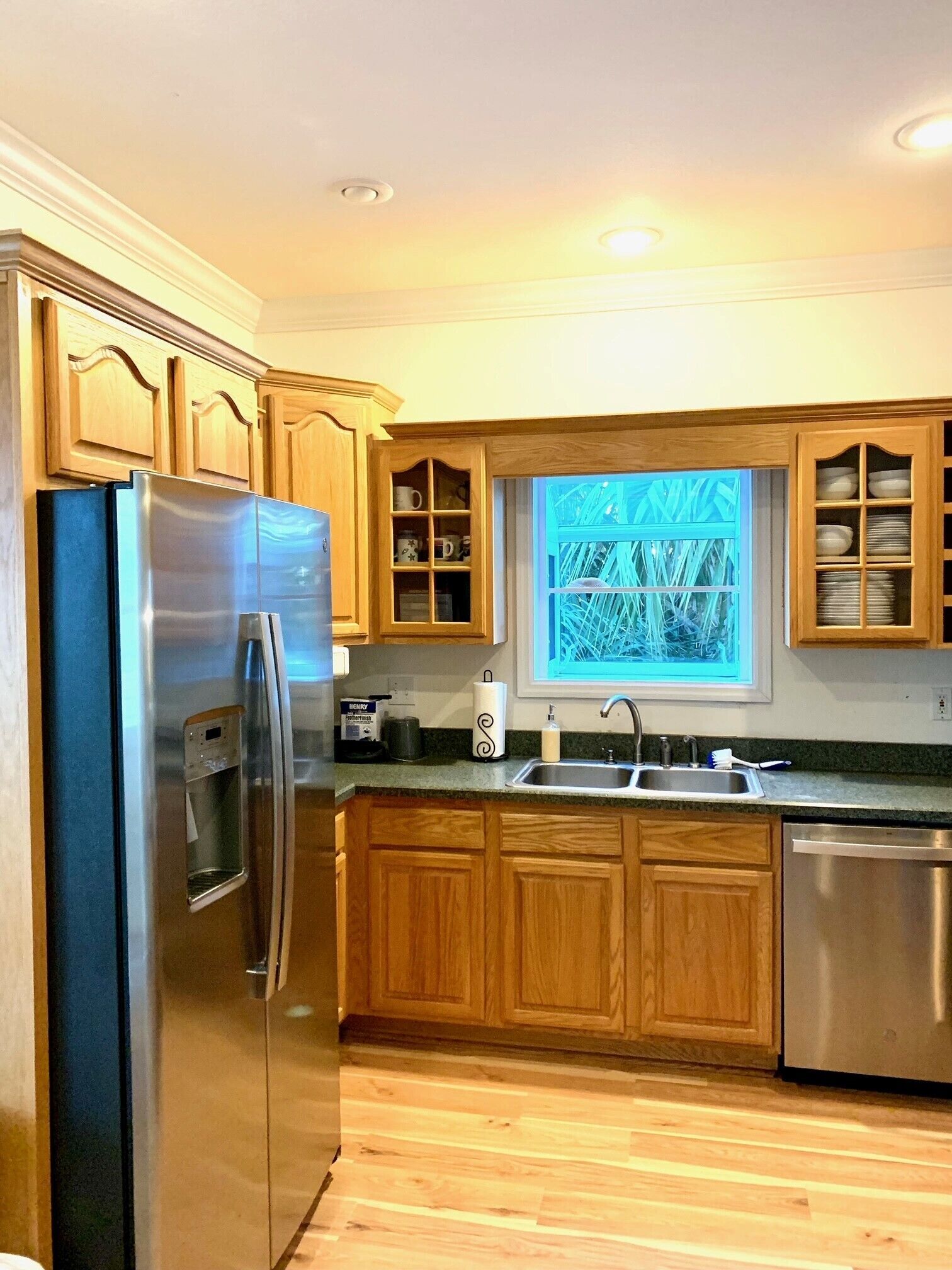 A kitchen with wooden cabinets, a stainless steel refrigerator, a built-in oven, a sink, and a window with a view of greenery.