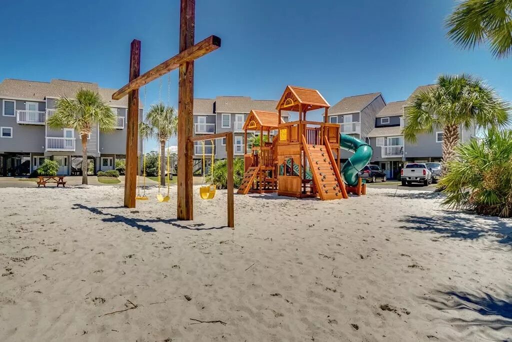 Une aire de jeux de plage avec un toboggan en bois et des structures d’escalade, entourée de palmiers et bordée, au loin, par une rangée de maisons de ville.