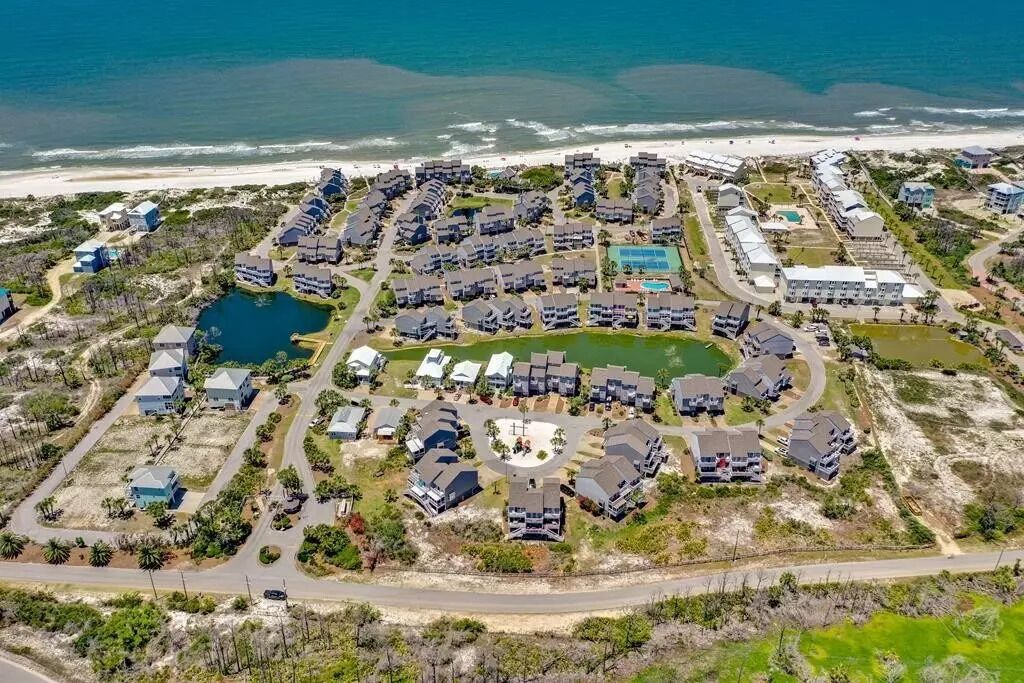 Vue aérienne d’un quartier résidentiel situé sur la côte, avec plusieurs bâtiments, une piscine centrale et une plage en arrière-plan.
