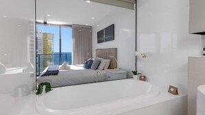 A modern bedroom with a large bed, a bedside table, a nightstand, a large mirror, and a view of a balcony with a cityscape. - Stunning Ocean Views Oracle Level 30 Parking (Broadbeach)