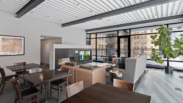A modern interior with a dining area, seating, a fireplace, and large windows offering a view of buildings outside.