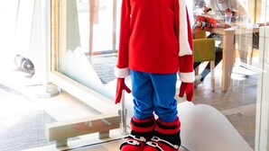 A mascot standing in a modern interior with glass doors, a fan, and a person seated at a table.