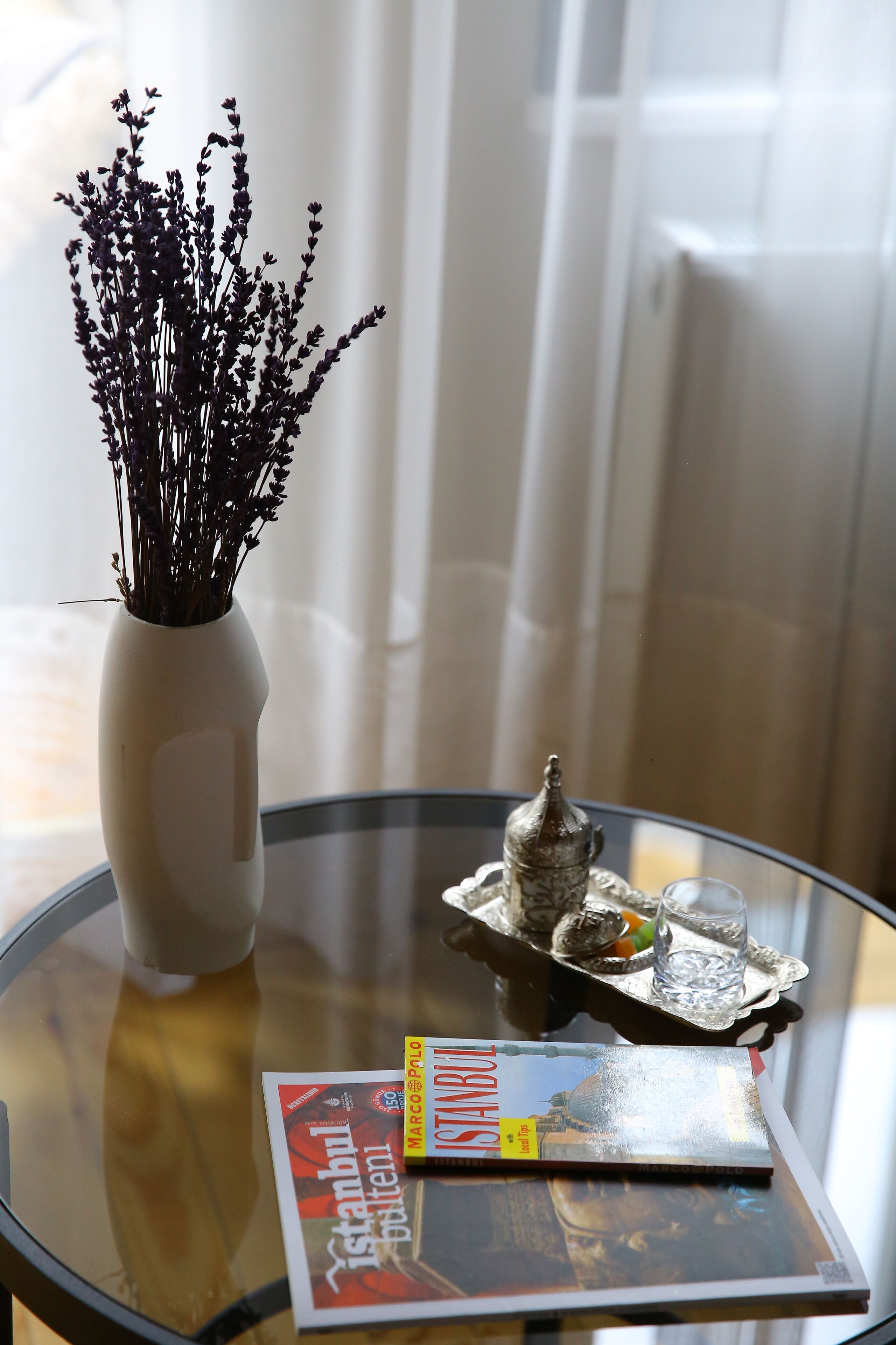 A round glass table with a vase of dried flowers, a tray with a decorative item and glasses, and a magazine titled "Istanbul."