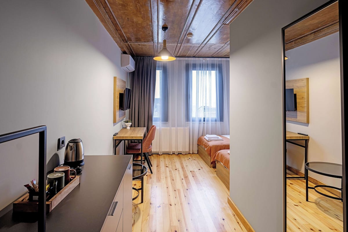 A compact hotel room with a wooden floor, a small desk with a chair, a bed with a headboard, a television mounted on the wall, and a window with sheer curtains.