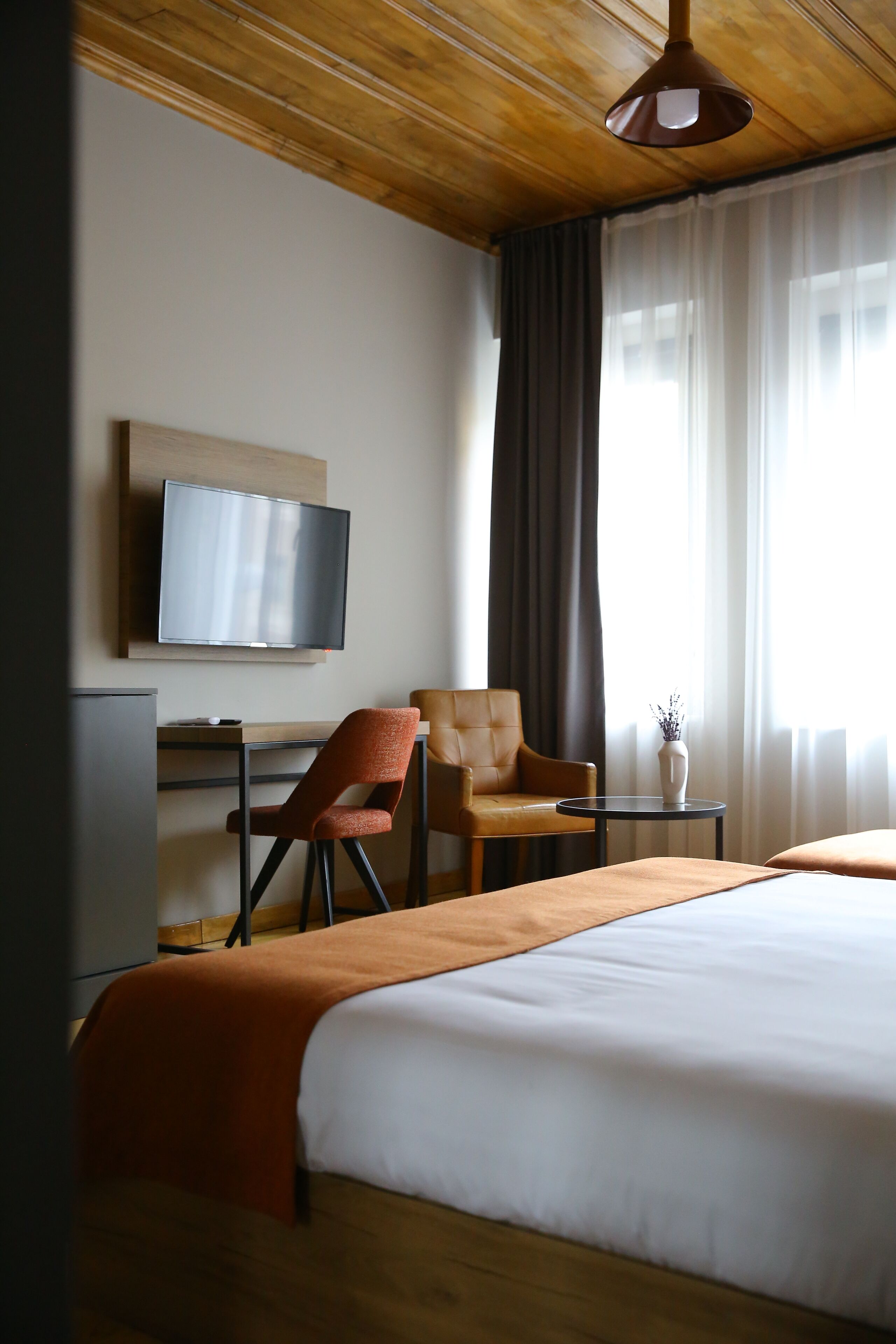 Habitación de hotel con cama, escritorio con televisor, dos sillones, una mesita con un florero y una ventana con cortinas.