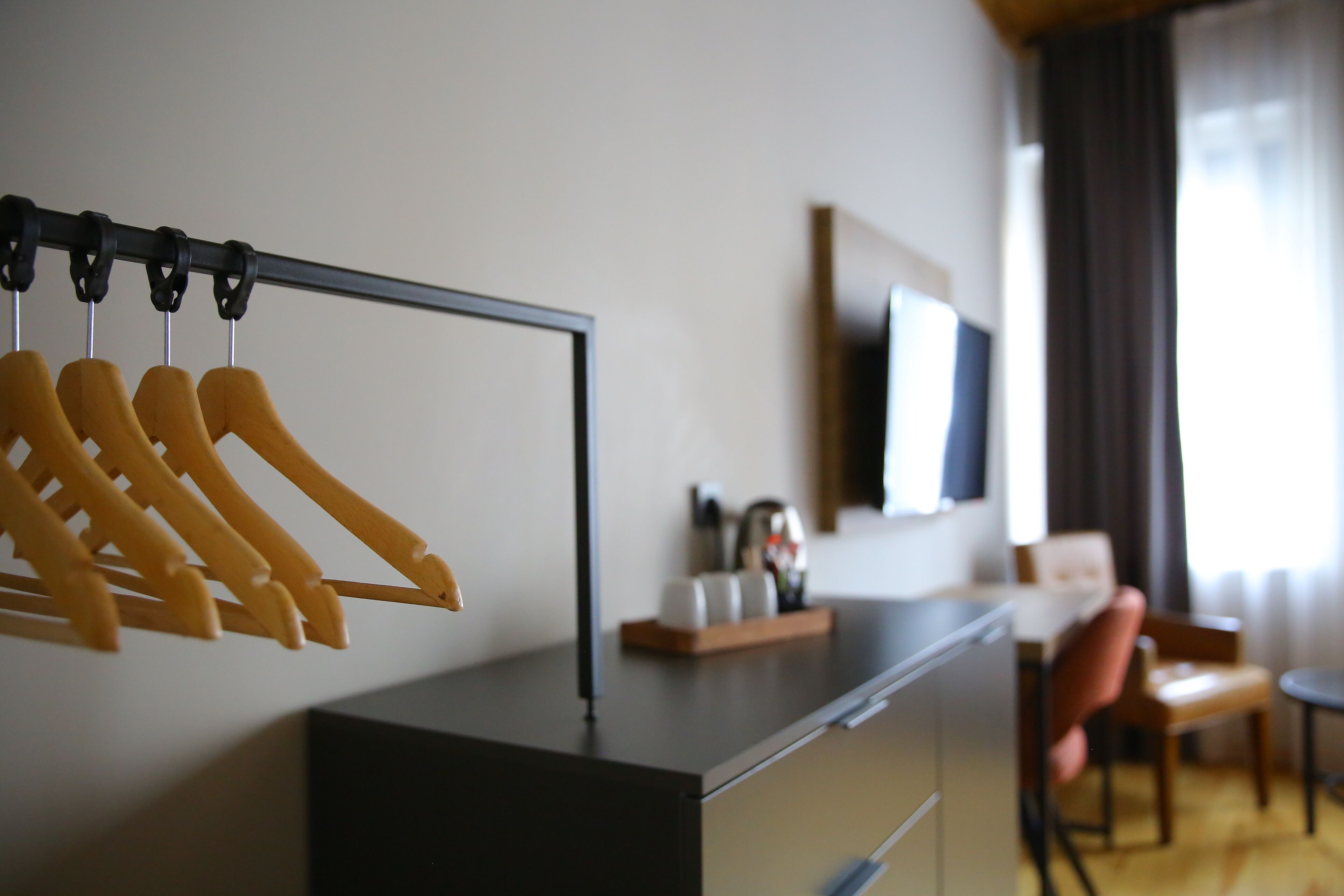 Hotellihuone, jossa on puinen naulakko, seinälle asennettu taulutelevisio, pieni pöytä, jossa on koriste-esine, tuoli ja ikkuna, jossa on verhot.