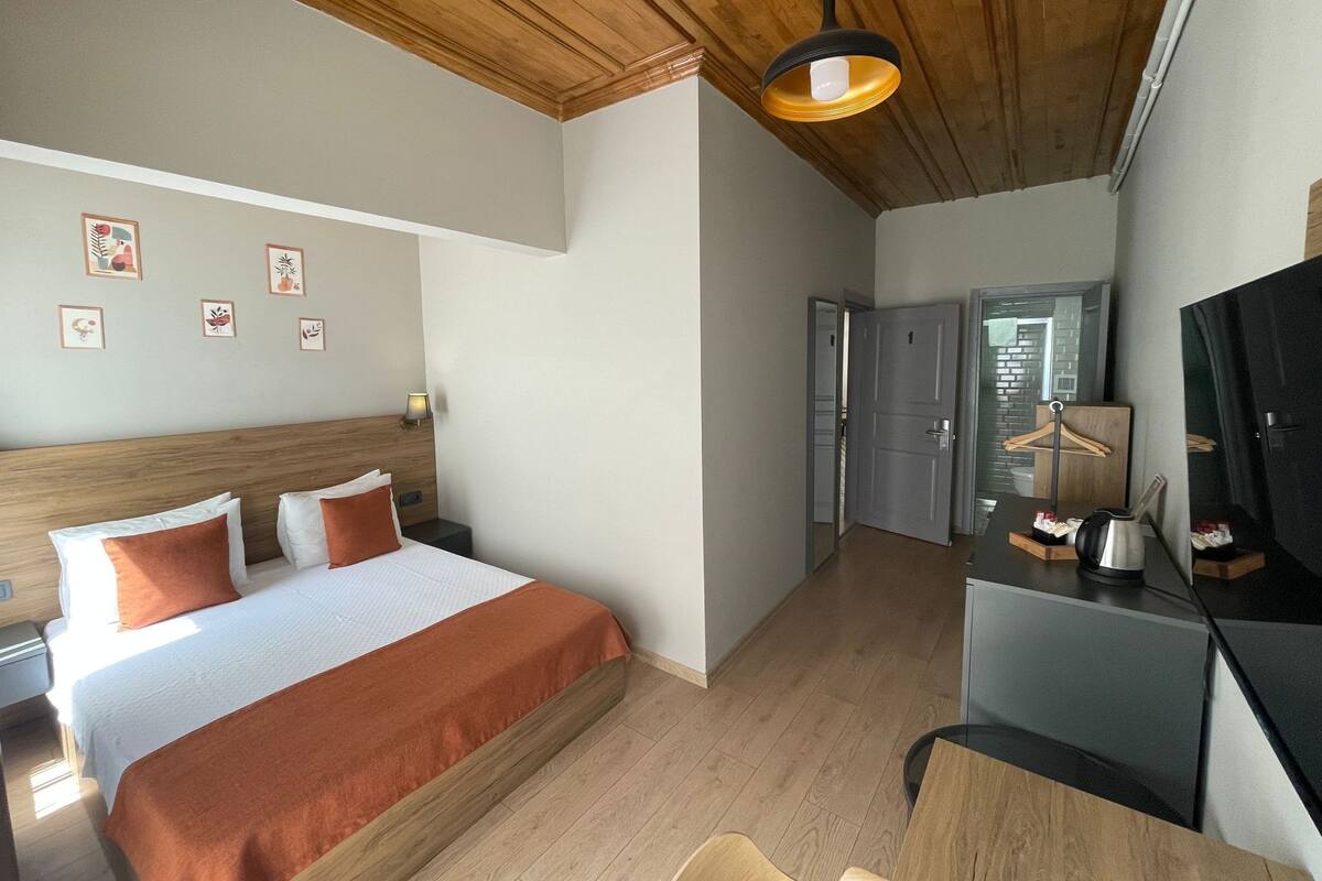 A hotel room with a wooden ceiling, a bed with white and orange bedding, a desk with a chair, and a TV.