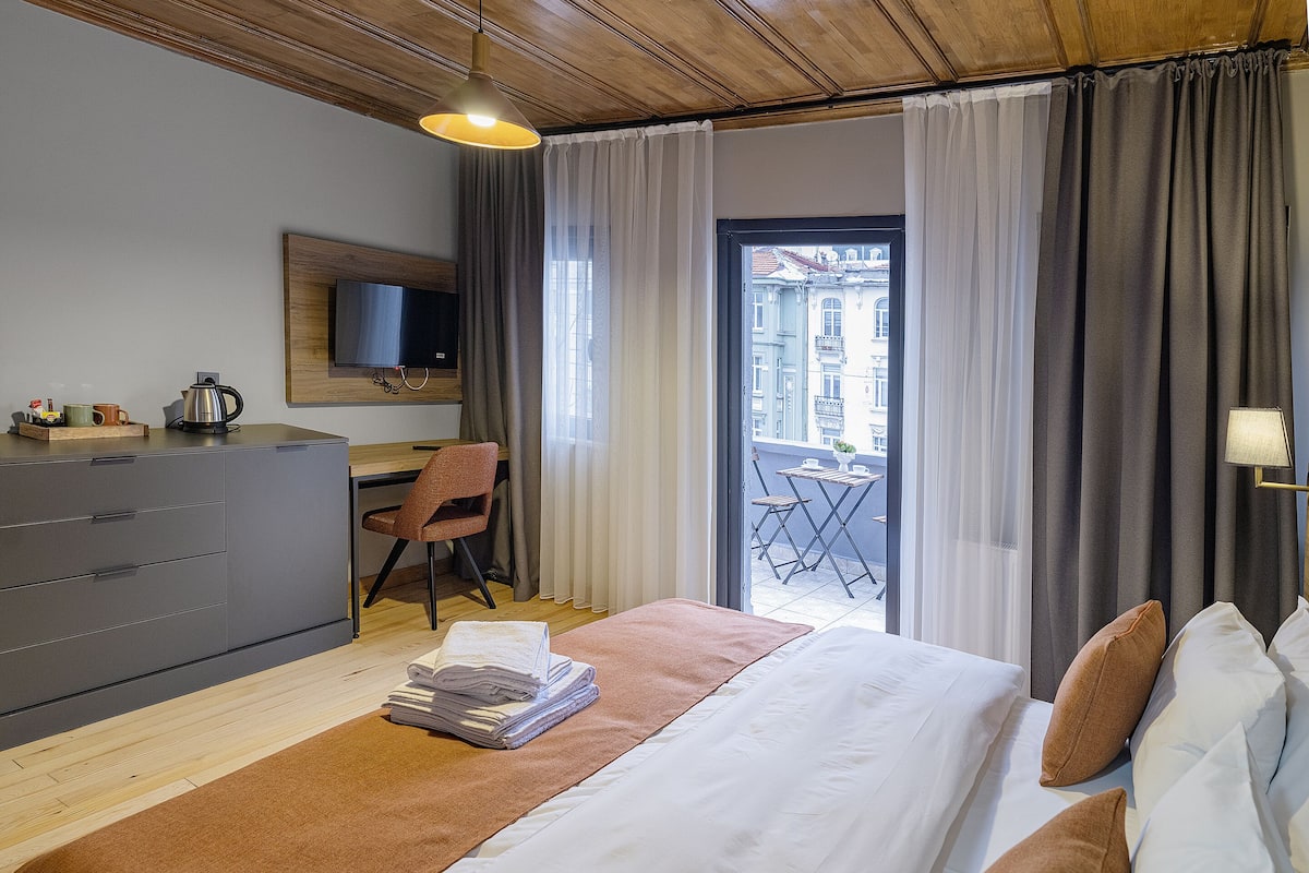 A hotel room with a bed, a desk, a chair, a TV, and a balcony with curtains.