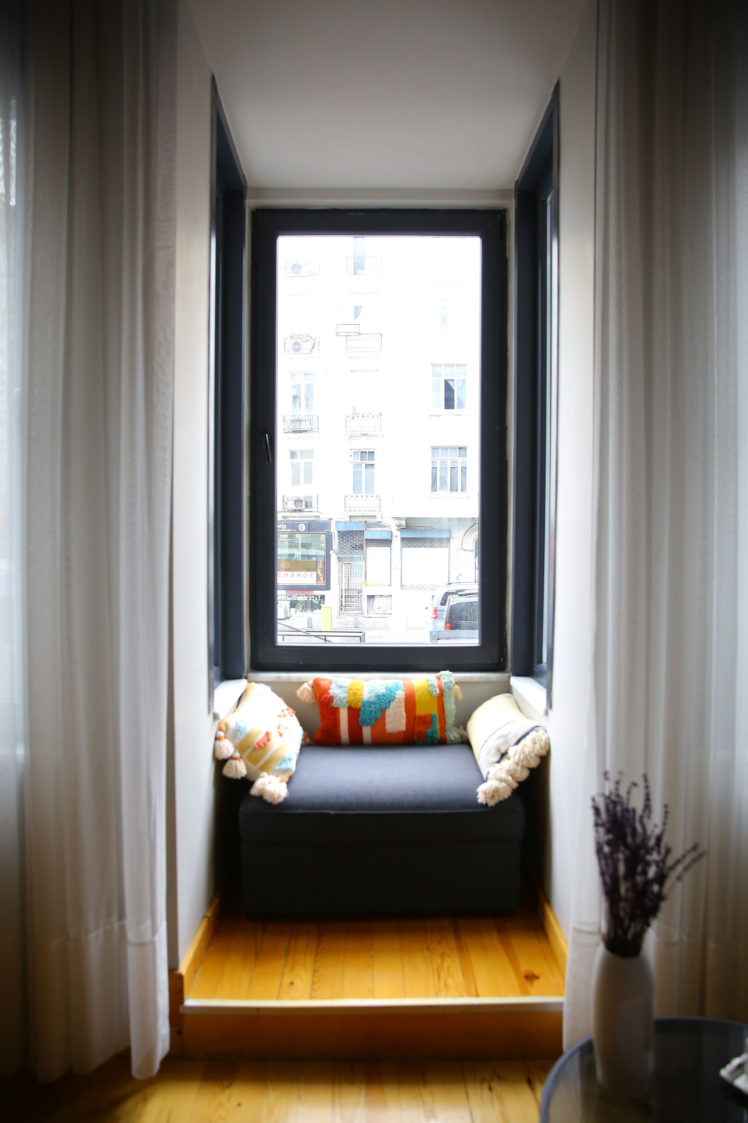 A room with a window seat, a vase with dried flowers, and a view of a building outside.