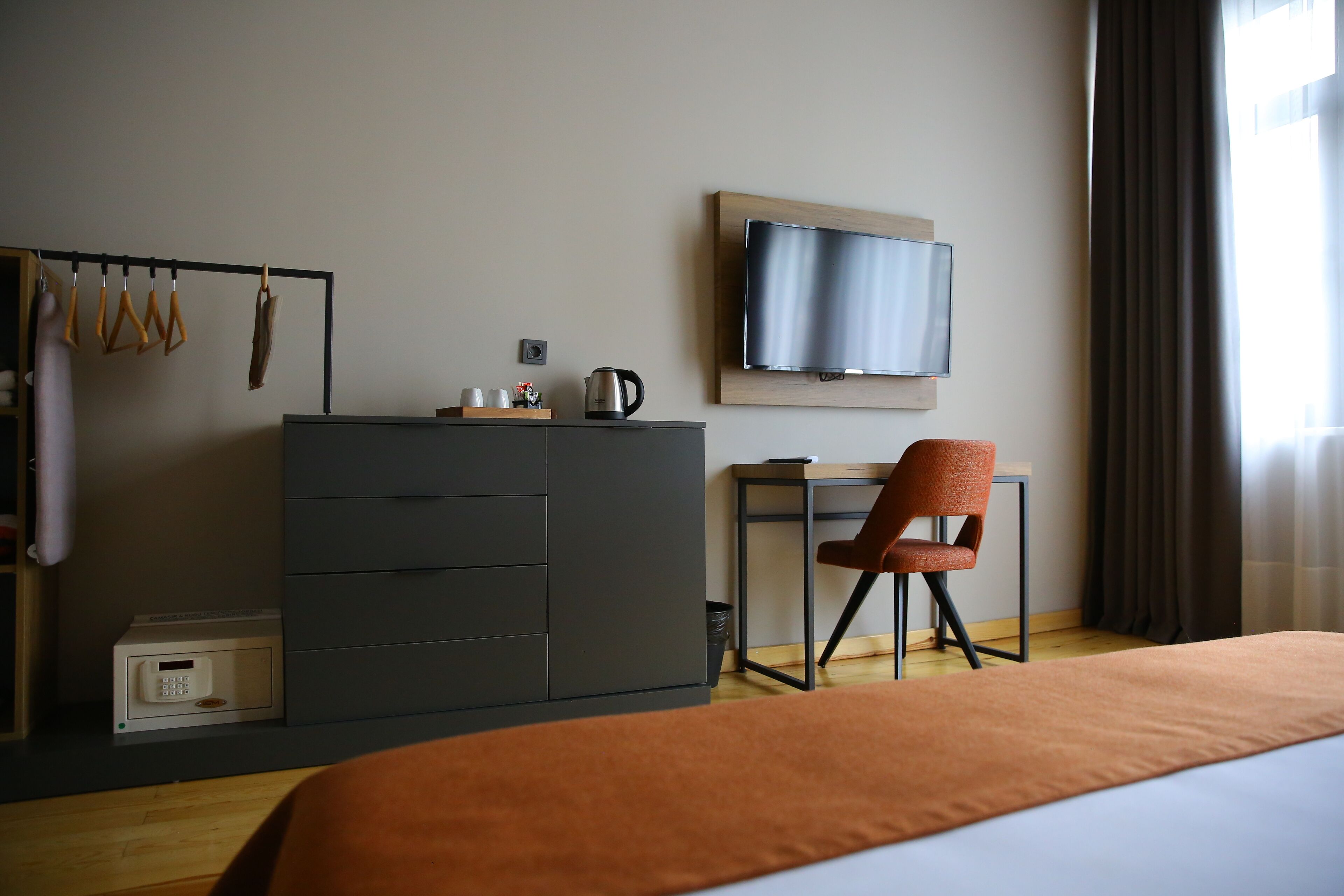 A hotel room with a bed, a desk, a chair, a television, a wardrobe, and a coat rack with clothes.