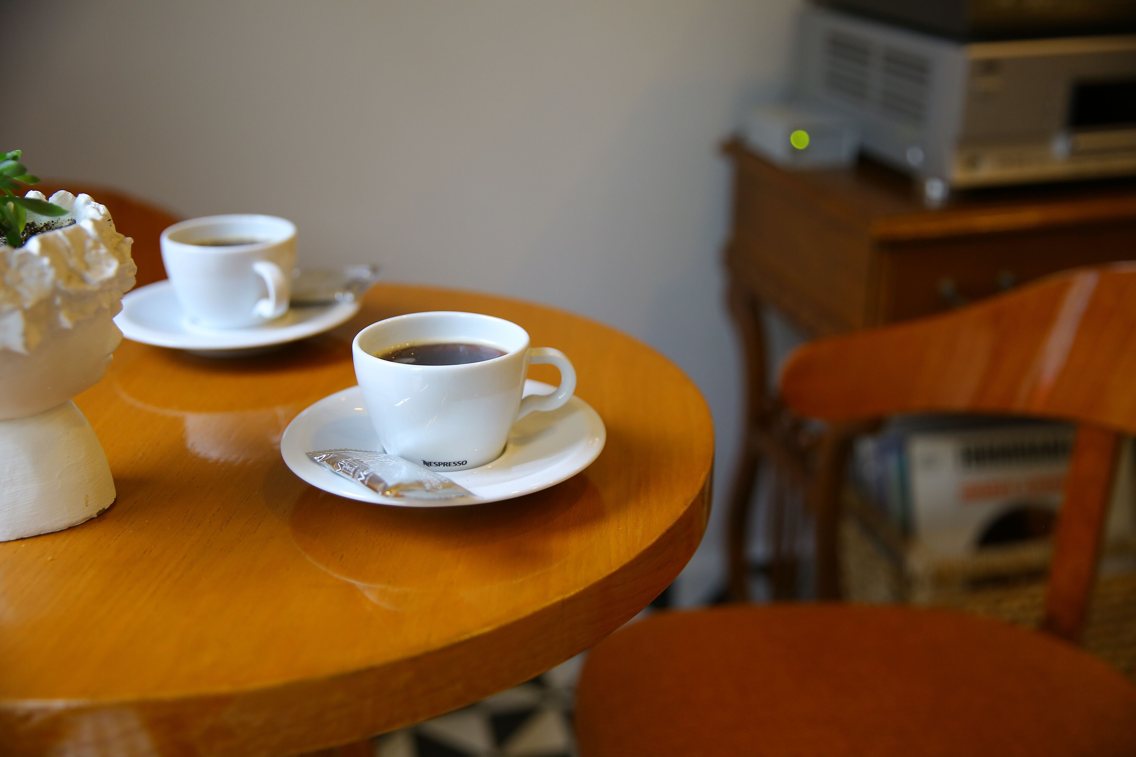 A wooden table with two white cups of coffee and a small white vase with a flower.