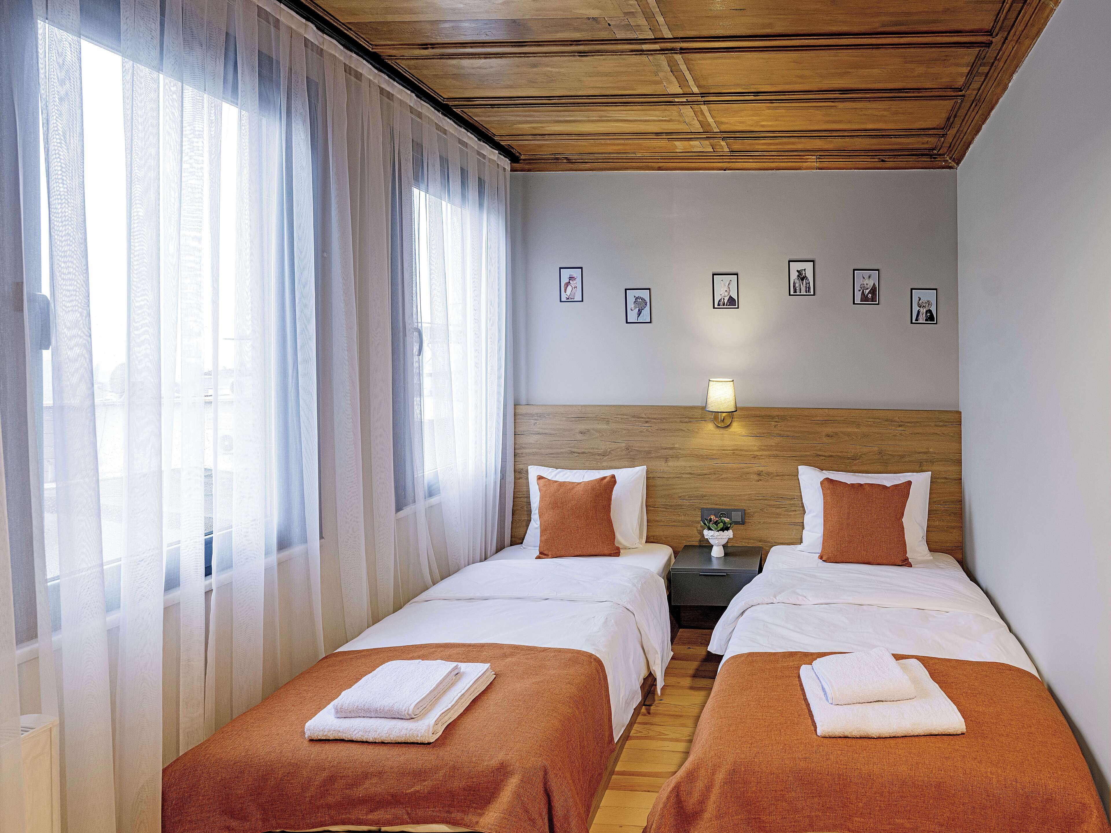 A hotel room with two beds, a wooden headboard, a nightstand with a lamp, and a window with sheer curtains.