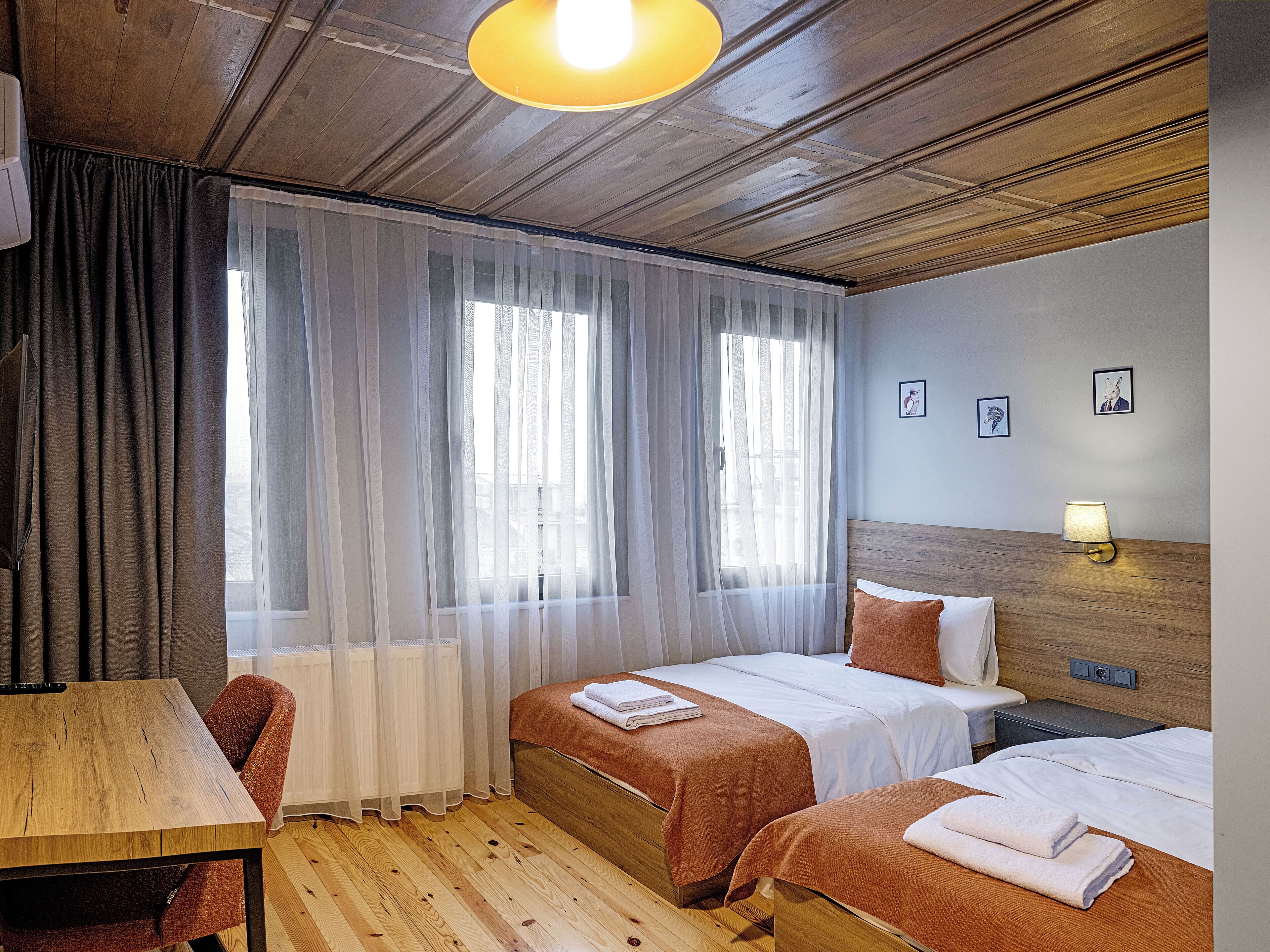 A hotel room with two beds, a wooden table, a chair, a desk, and a window with sheer curtains.