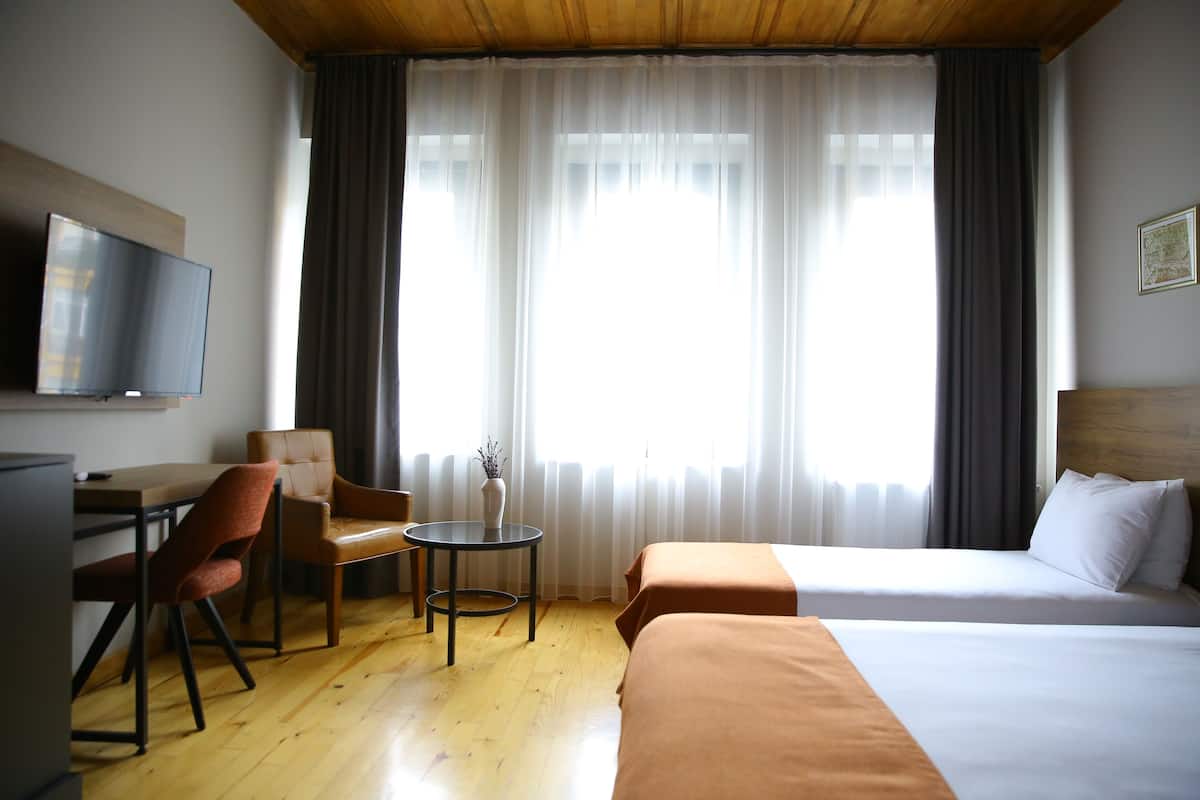 A hotel room with a wooden ceiling, a TV mounted on the wall, a desk with a chair, two beds with white bedding, and a large window with sheer curtains.