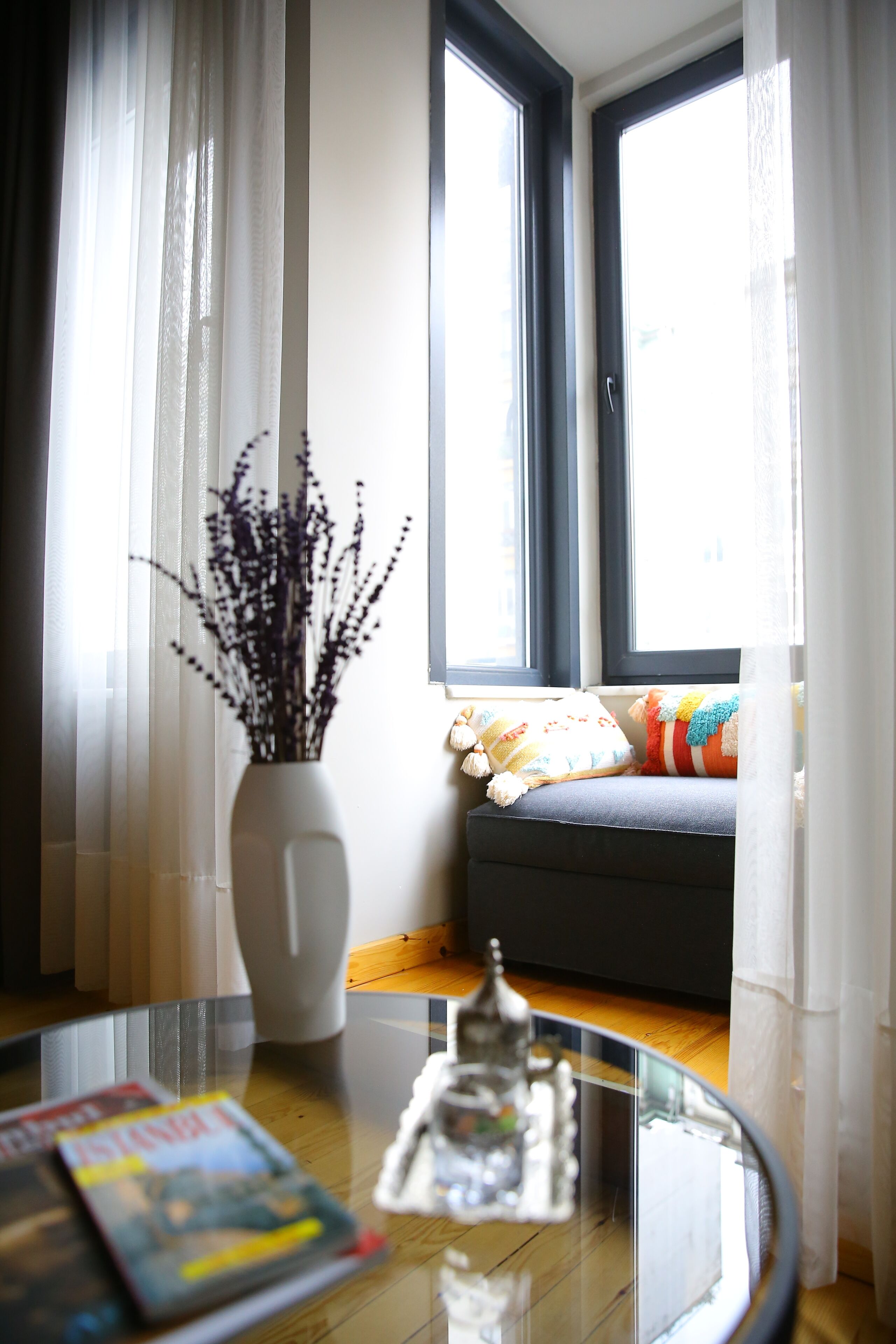 Ein Zimmer mit Fenster, einer Couch mit bunten Kissen, einer weißen Vase mit Trockenpflanzen und einem Glastisch mit Zeitschriften und einem dekorativen Gegenstand.