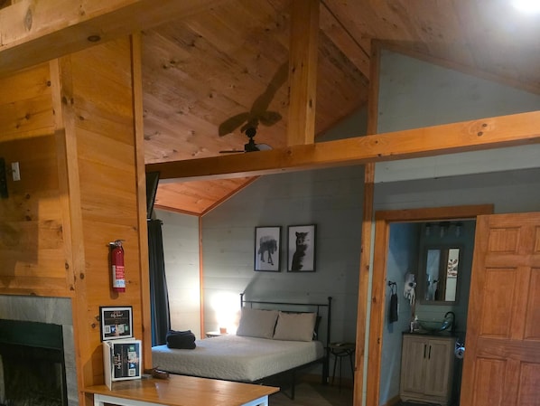 Ein Interieur einer Holzhütte mit Bett, Deckenventilator, Kamin und einem Badezimmer mit Spiegel und Waschbecken.