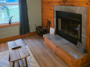 Ein gemütliches Zimmer mit Kamin, einer hölzernen Bank, einem Tisch und einem Fenster mit Blick ins Freie.