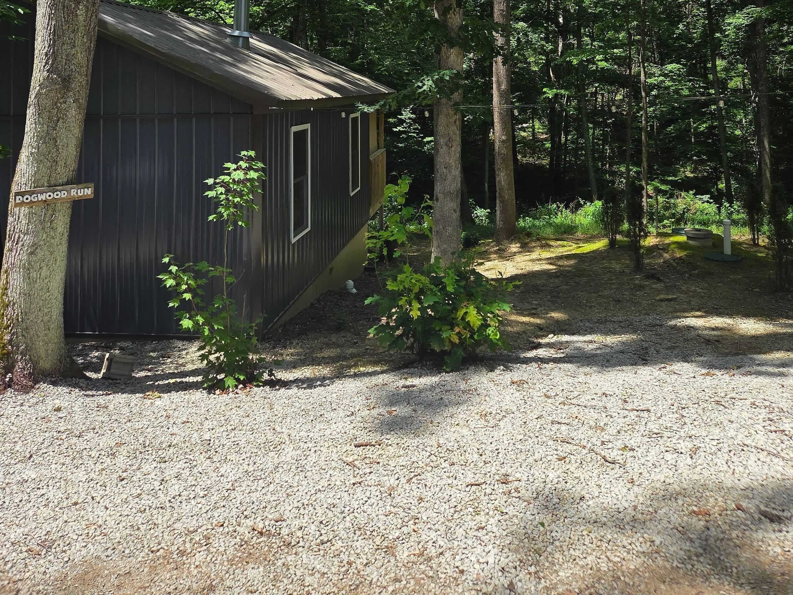 A small, dark wooden cabin with a gravel ground, surrounded by trees.
