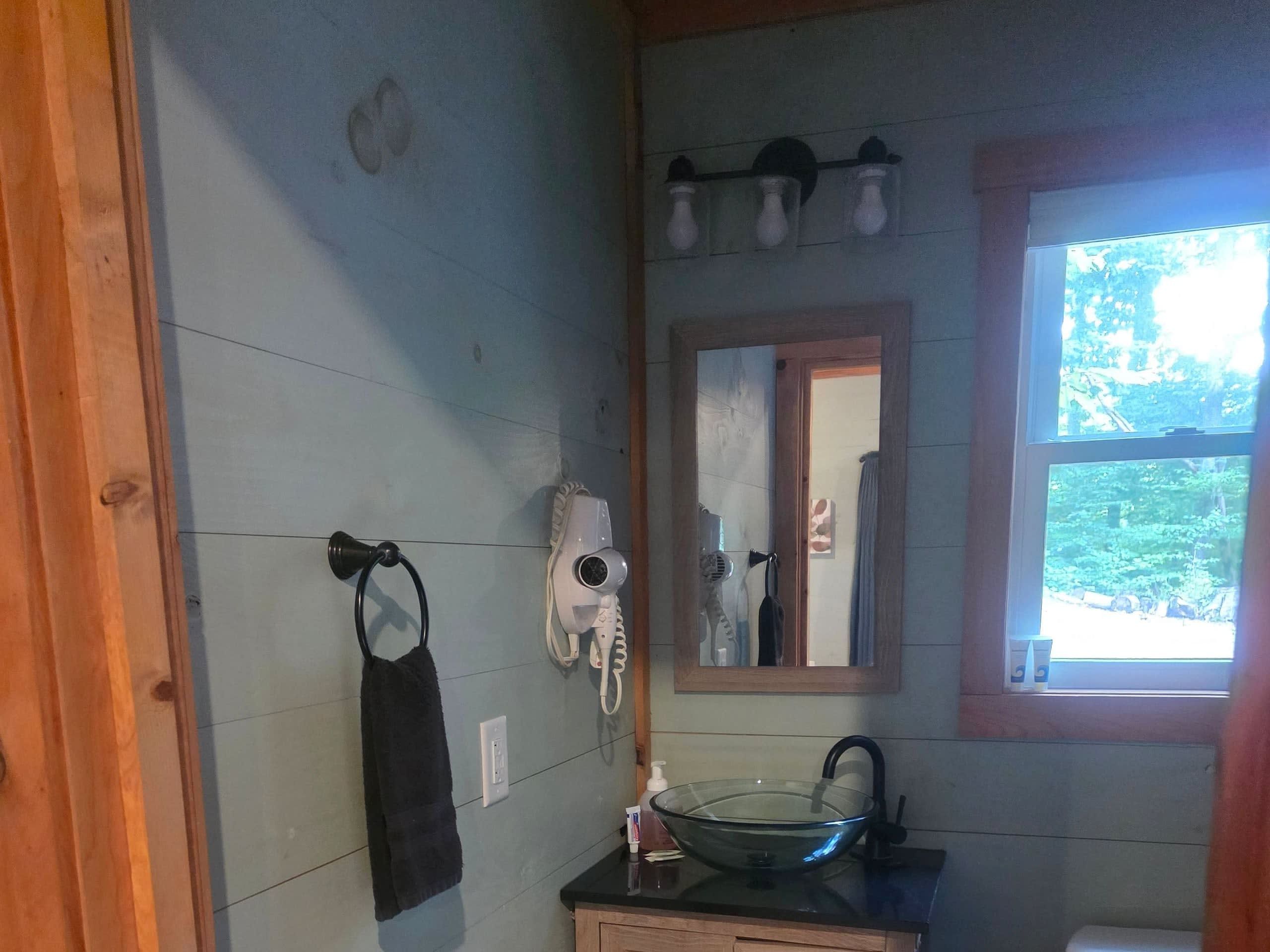 Ein Badezimmer mit einem Waschbecken aus Glas, einem Handtuch auf einem Halter, einem Fön und einem Spiegel über dem Waschbecken.