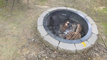 A circular stone fire pit with logs and ash, surrounded by a grassy area with a metal bench in the background.