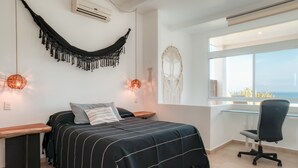 Een slaapkamer met een bed, nachtkastjes, een bureau, een stoel, een hangmat, macramewanddecoratie en een groot raam met uitzicht.
