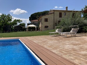 Una piscina con cubierta de madera, dos sillas reclinables y un edificio de piedra al fondo.