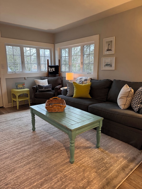 Un salon chaleureux avec une table basse verte, un canapé foncé agrémenté de coussins à motifs, un panier en osier et un fauteuil avec un coussin orné de la mention « Bienvenue ».