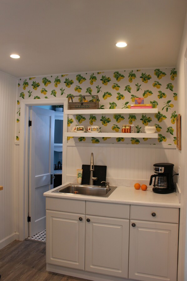 A kitchen with white cabinets, a stainless steel sink, and a coffee maker. There are two oranges on the counter. - Cozy Cottage (Ludington)