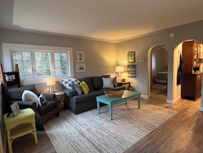 A living room with a sofa, a coffee table, a lamp, and a small side table. There are framed pictures on the wall and a window with a view of trees.