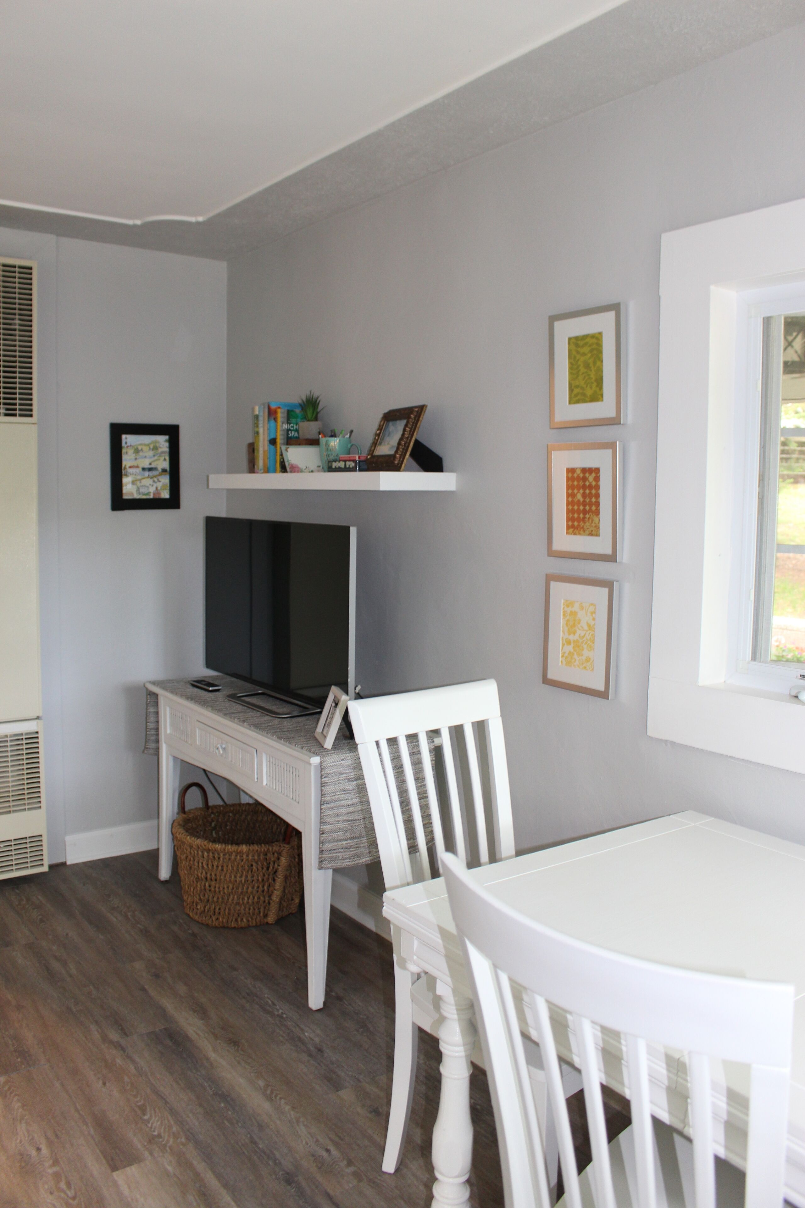 A room with a white dining table, a white chair, a television on a white stand, a wicker basket, and framed artwork on the wall.