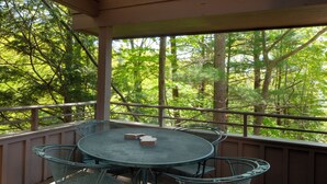 A covered outdoor area with a round metal table and chairs, surrounded by wooden decking and a view of a forested area. - Hawk's Nest 5 (Glen Arbor)
