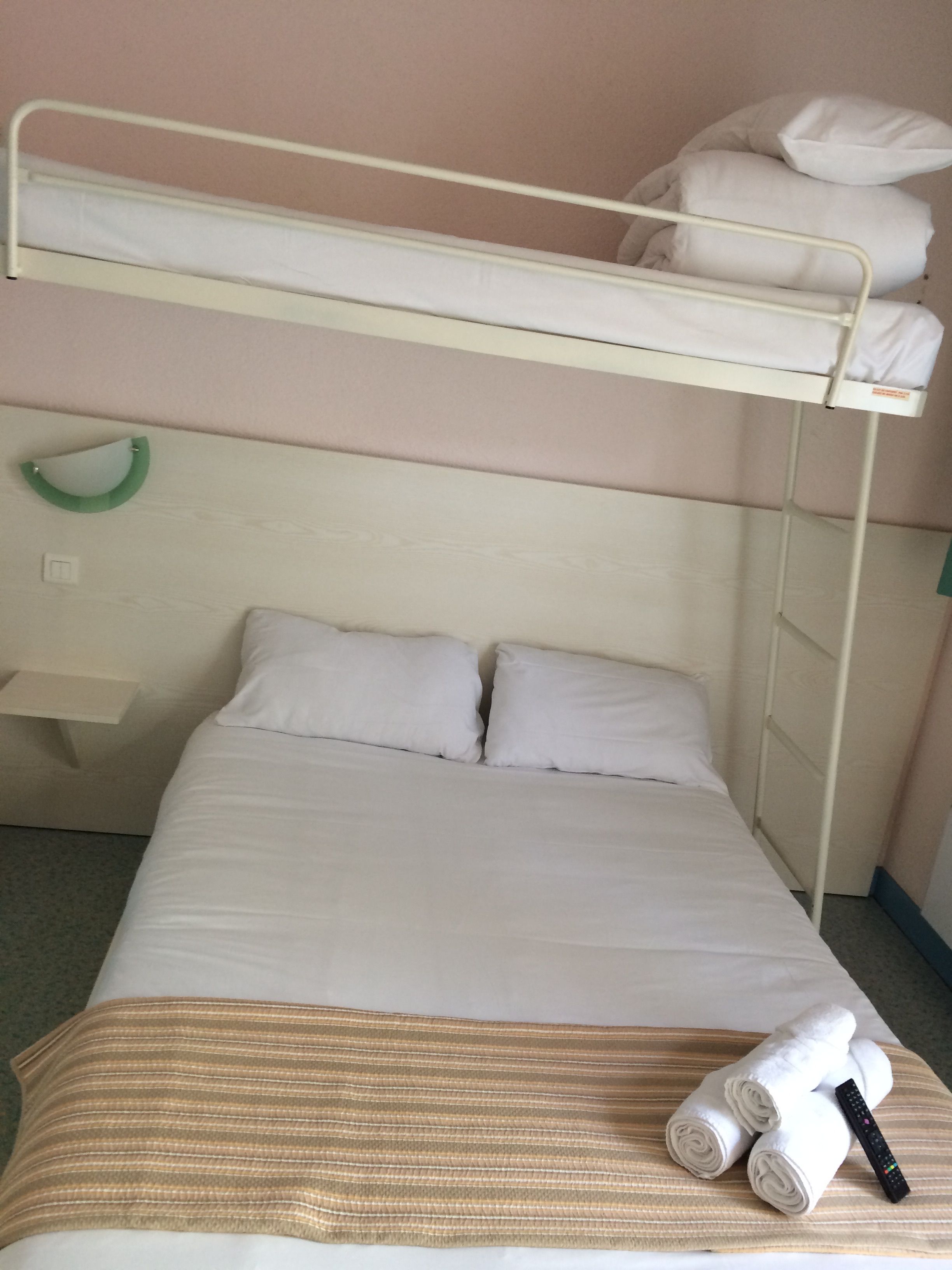 Uma cama beliche com uma cama inferior e um beliche superior. Há almofadas na cama de baixo e um objeto verde fixado na parede à esquerda.