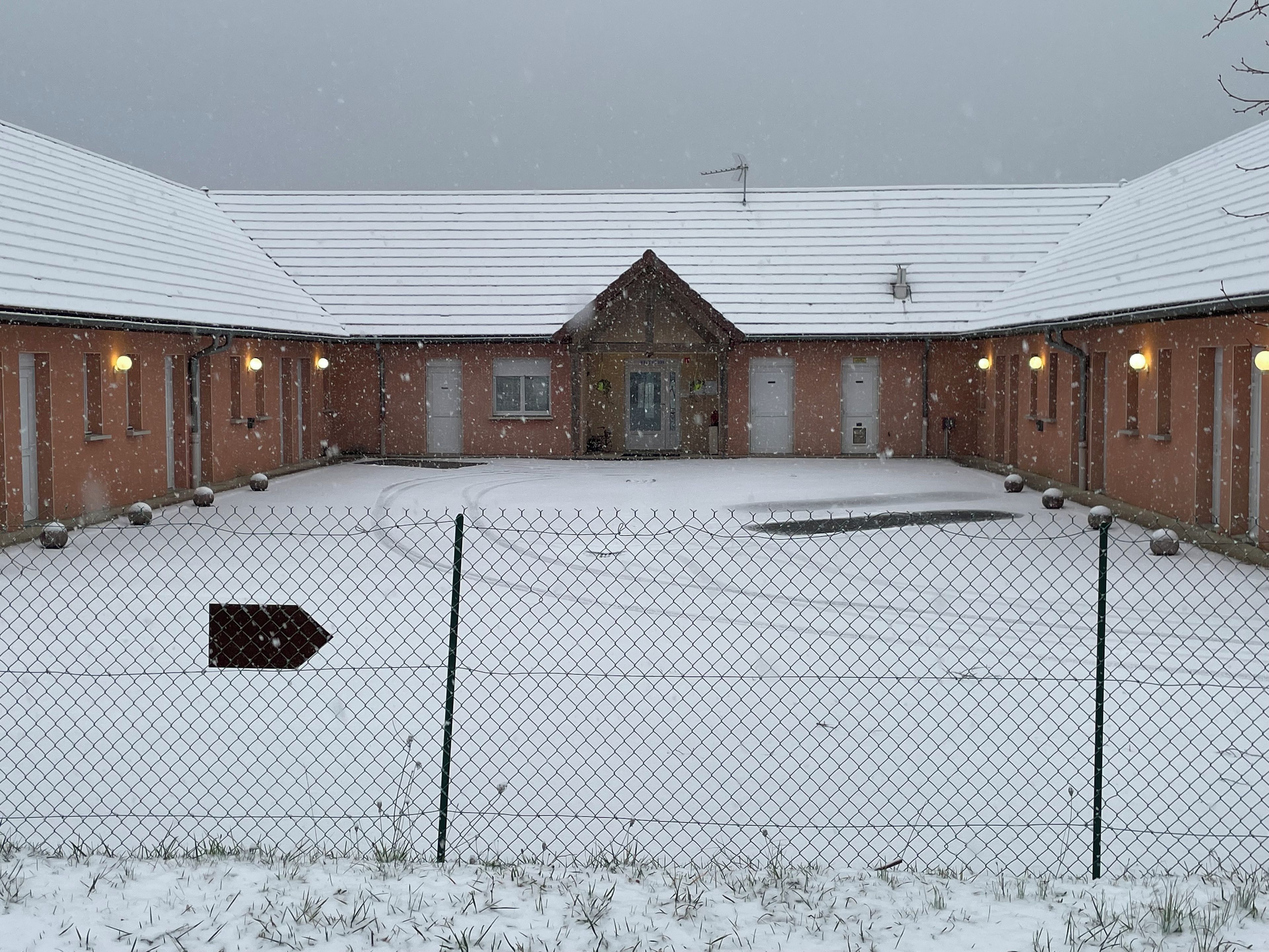 A fenced courtyard with a building featuring multiple doors and windows, surrounded by snow-covered ground.