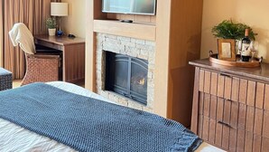 Habitación de hotel con chimenea, televisor montado encima, escritorio con lámpara y cama con manta azul texturizada.