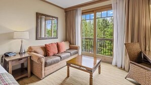 Habitación de hotel con sofá, una mesita de centro de madera, lámpara, escritorio y silla. Cuenta con un ventanal con cortinas y vistas a árboles.
