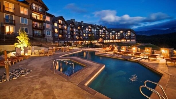 Un hotel con piscina, mobiliario exterior y montañas al fondo.