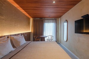 A hotel room with a large bed, a small table with chairs, a TV mounted on the wall, and a window with curtains.