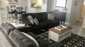 Een moderne woonkamer met een donkergrijze bank, een salontafel en een kamerkleed. Er is een eethoek met een tafel en stoelen, en een keuken op de achtergrond.