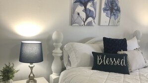 Een slaapkamer met een wit bed, een nachtkastje met een lamp en twee bloemrijke wanddecoraties.