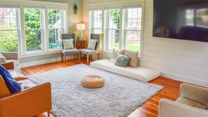 A living room with a large window, a sofa, a chair, a TV, and a lamp. - Beautiful and pristine 4 bedroom home,  7 minute walk to the Beach! (West Yarmouth)