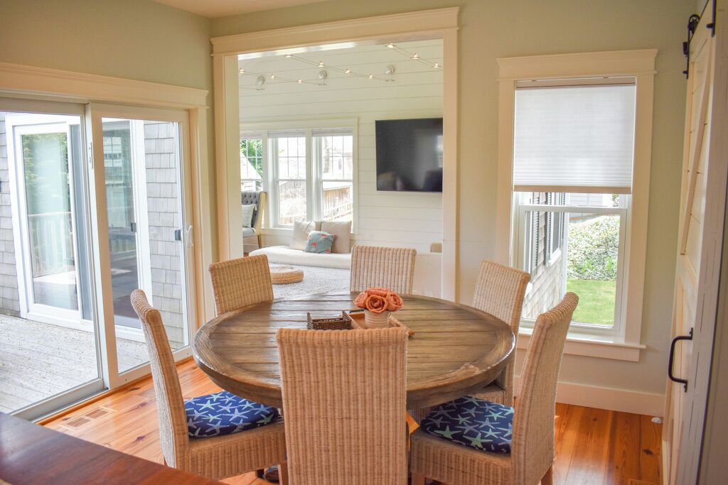 A dining area with a round table and wicker chairs, large windows, and a television on the wall.