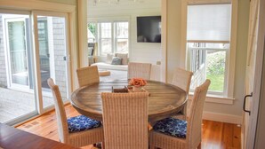 A dining area with a round table and wicker chairs, large windows, and a television on the wall. - Beautiful and pristine 4 bedroom home,  7 minute walk to the Beach! (West Yarmouth)