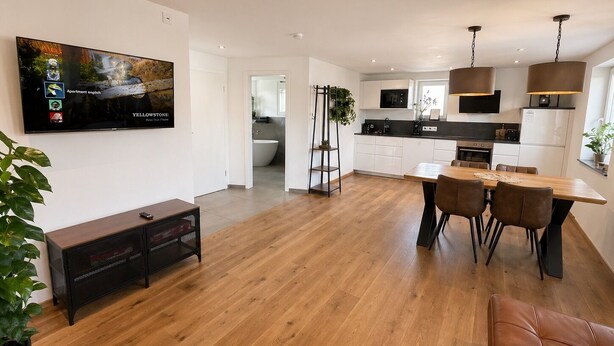 Une cuisine moderne avec des meubles blancs, un coin repas avec une table et des chaises en bois, et un téléviseur fixé au mur qui diffuse une publicité.